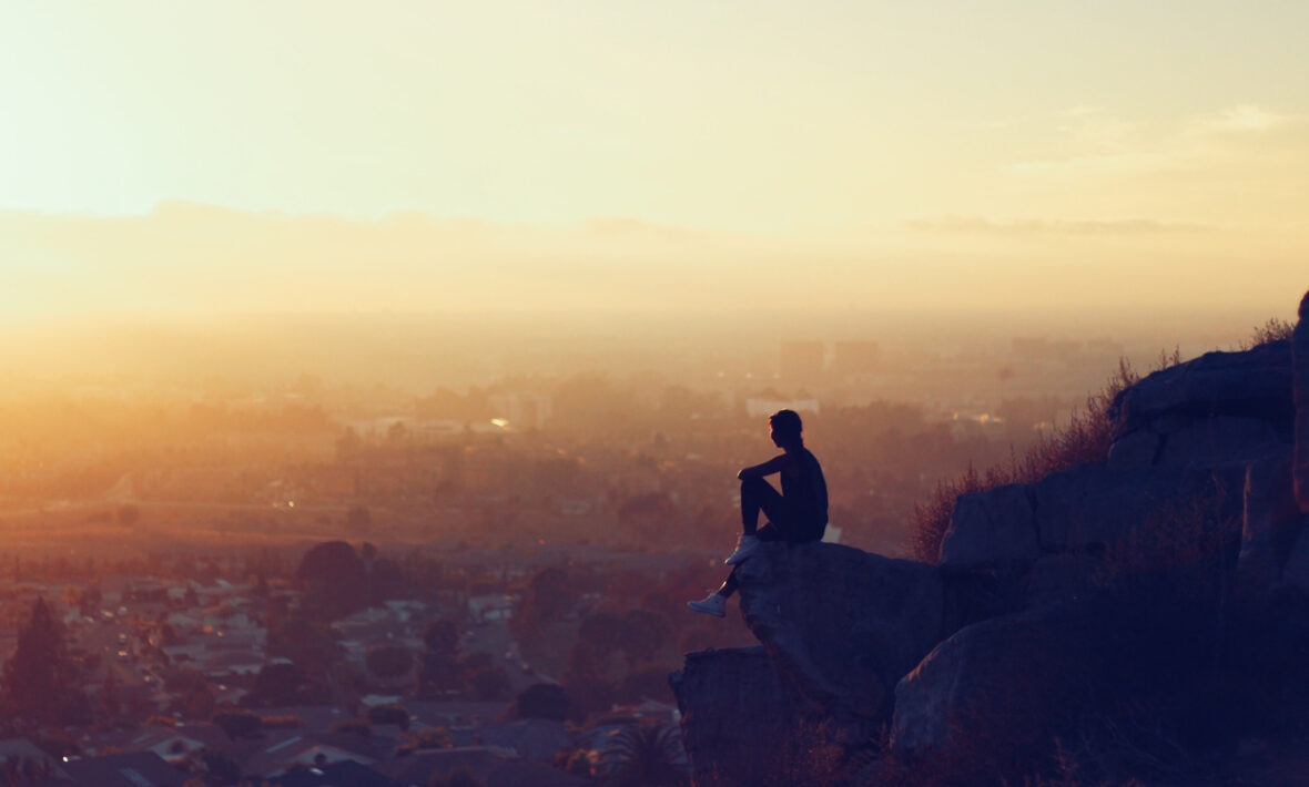 self improvement - image of a man sitting on a rock looking out on to a city at sunset