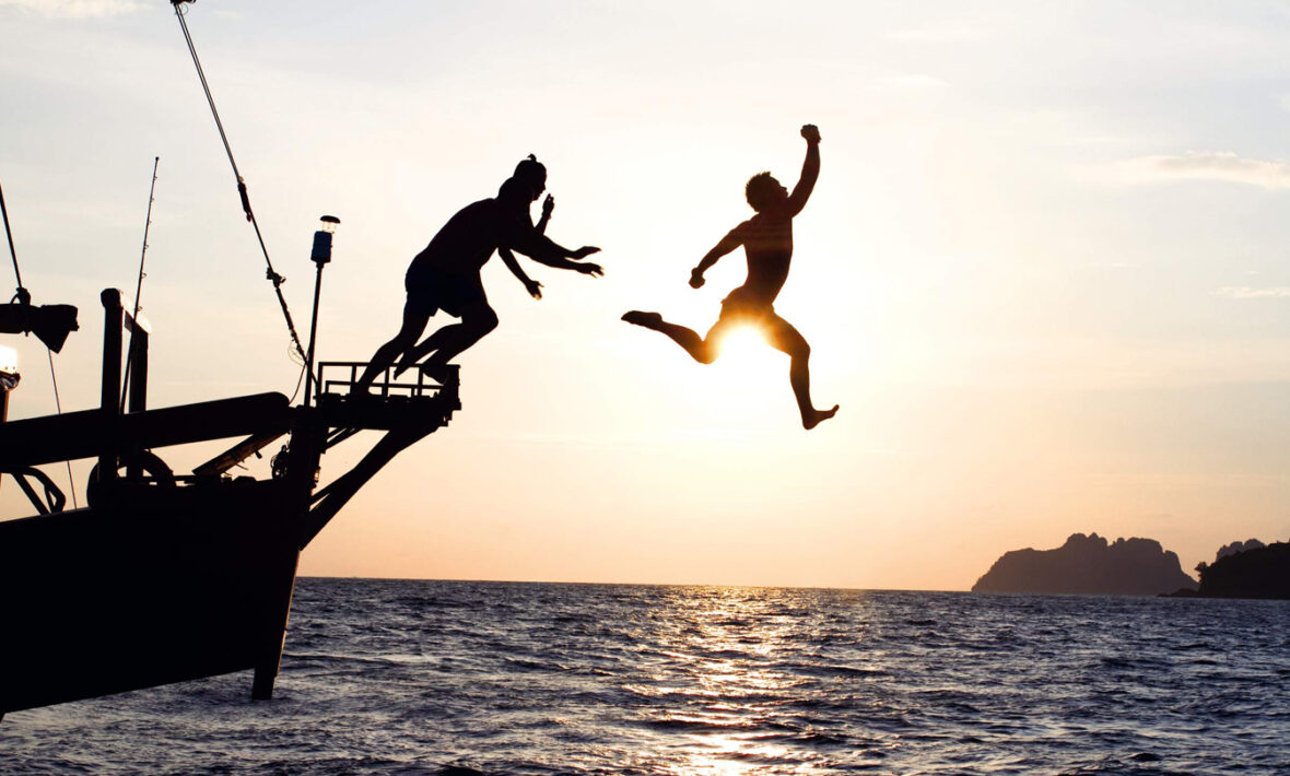 Two people jumping off a boat at sunset while capturing the moment in the best travel books.