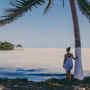 A woman is standing under a palm tree near the ocean, one of the best places to travel in November.