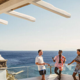 Three men standing on a balcony overlooking the ocean in Greece.