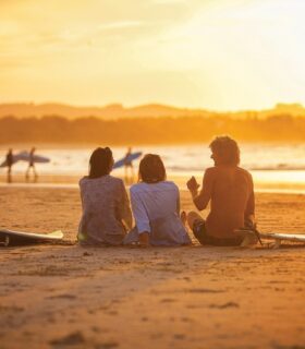 Friends sitting on a beach at sunset watching surfers at the water's edge.