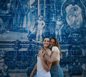 Two women hugging during their travel adventure in front of a blue tiled wall.