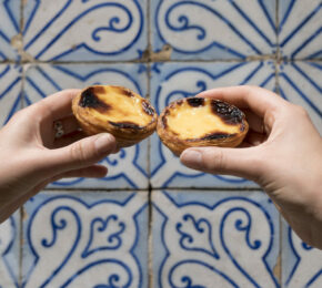 A pair of hands holding two pastries in front of a blue and white tiled wall in Portugal.