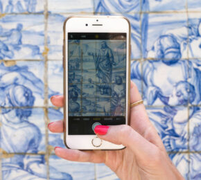 A person using travel apps on a cell phone in front of a blue tiled wall.