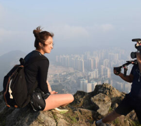 A woman, a travel filmmaker, sitting on top of a mountain with a camera.