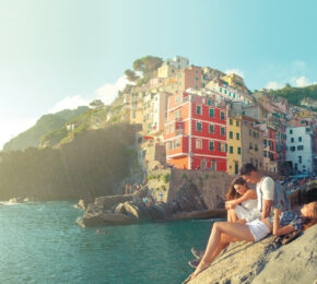 A couple is having a summer fling while sitting on a rock overlooking the water.