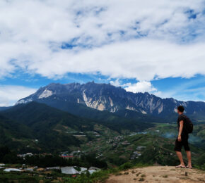 A man backpacking on a budget standing on top of a mountain overlooking a village.