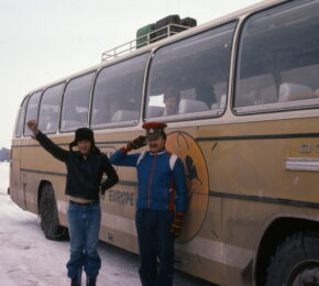 Two people standing in front of a retro yellow bus, reminiscent of a contiki ad.