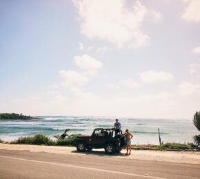 Two people enjoying one of the best road trips in the world standing on top of a jeep in front of the ocean.