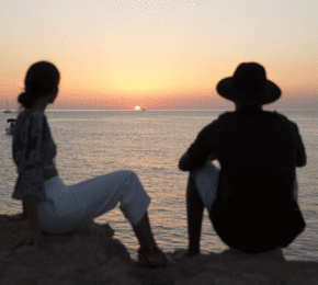 Two people experiencing true love while sitting on a cliff overlooking the ocean at sunset.