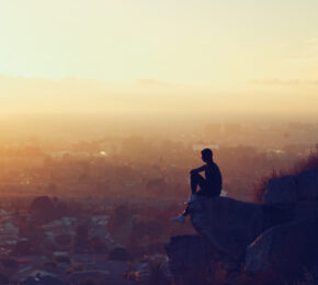 self improvement - image of a man sitting on a rock looking out on to a city at sunset