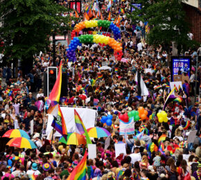 A crowd of people participating in a pride event, proudly walking down a street with rainbow flags.
