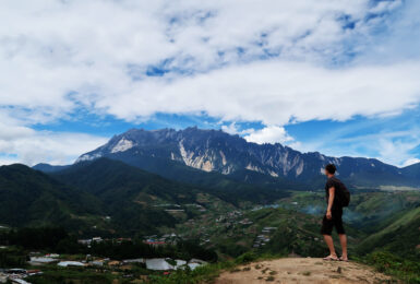 A man backpacking on a budget standing on top of a mountain overlooking a village.