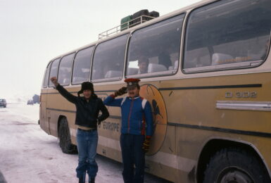 Two people standing in front of a retro yellow bus, reminiscent of a contiki ad.