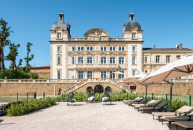 A large stone Contiki Château building is in the middle of a courtyard with lounge chairs and umbrellas.