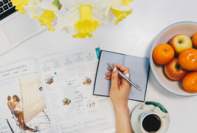 A woman is writing on a notebook while sitting at a desk with oranges and a laptop.