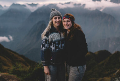 Two millennial women standing on top of a mountain with mountains in the background.