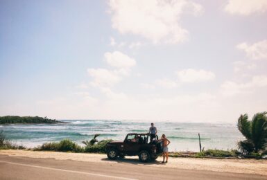 Two people enjoying one of the best road trips in the world standing on top of a jeep in front of the ocean.