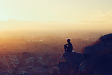 self improvement - image of a man sitting on a rock looking out on to a city at sunset