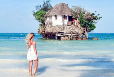 A woman is standing in front of a small house in the ocean, resembling a rock restaurant in Zanzibar.