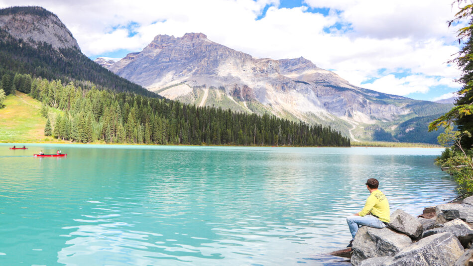 A person is sitting on rocks near a lake, contemplating their next travel adventure based on their zodiac sign.