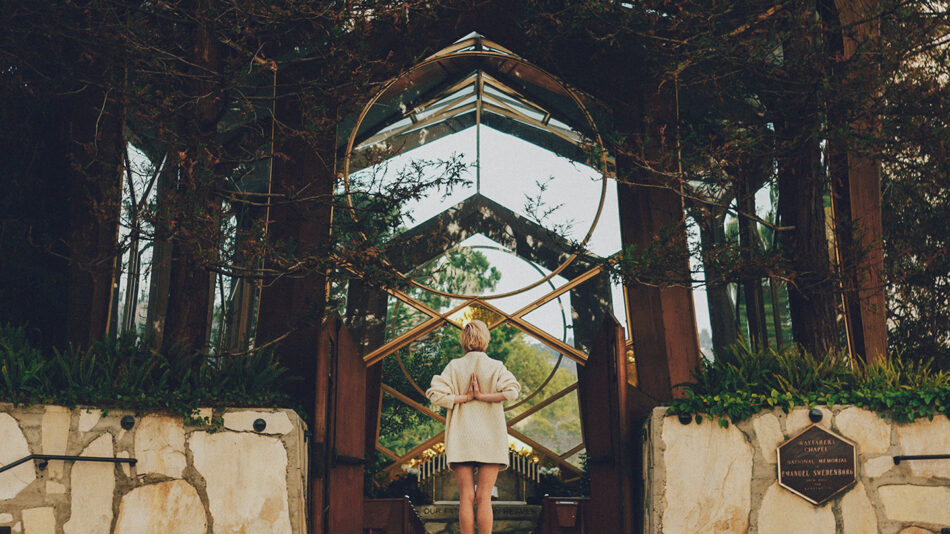 A woman practicing yoga in a white dress, peacefully positioned in front of a glass door.