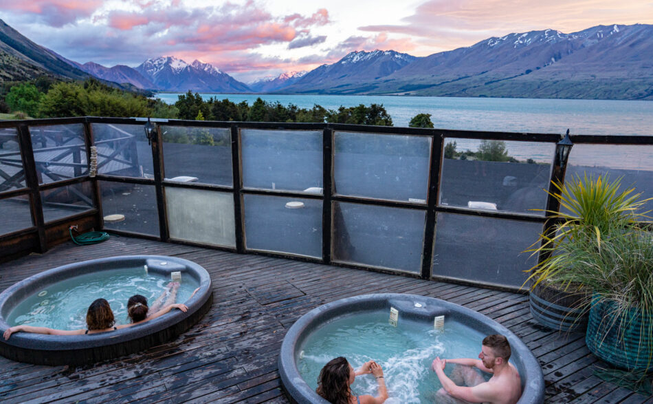 Three people sitting in hot tubs on a deck overlooking lake wanaka.