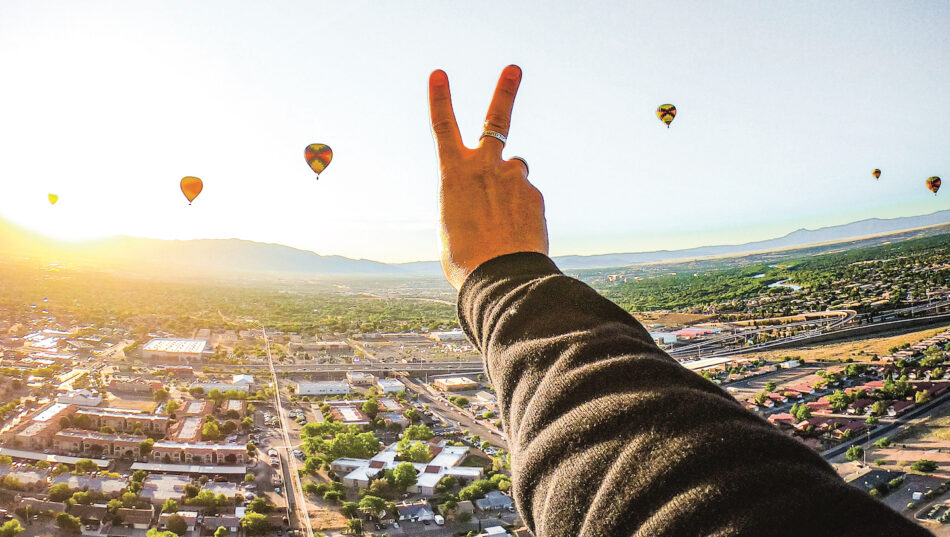 A person making a peace sign over a city with hot air balloons, symbolizing how travel can change you.