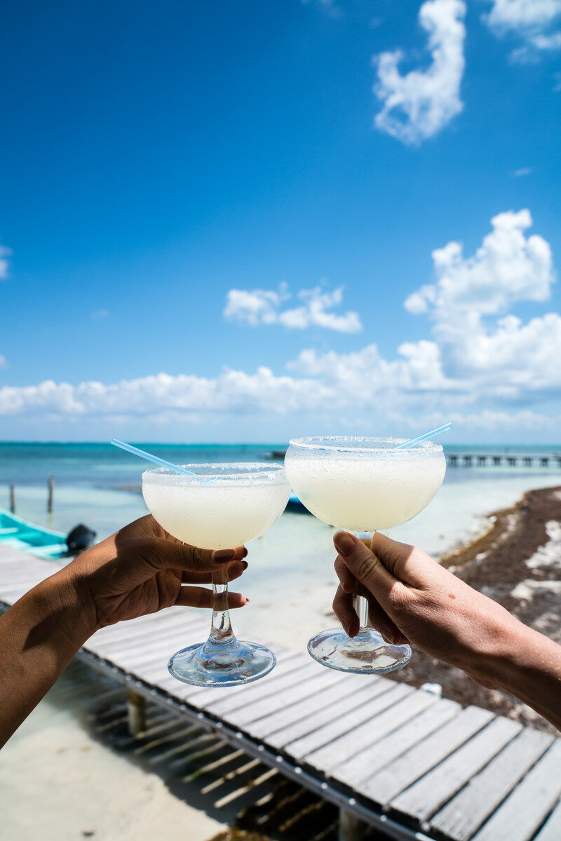 Two LGBTQ individuals celebrating with margaritas on a dock in Mexico.