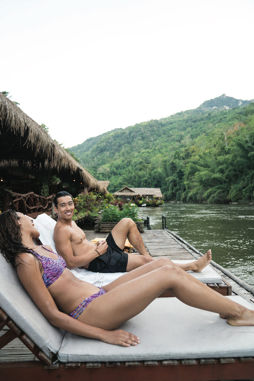 A man and woman relaxing on a wooden deck next to a river.