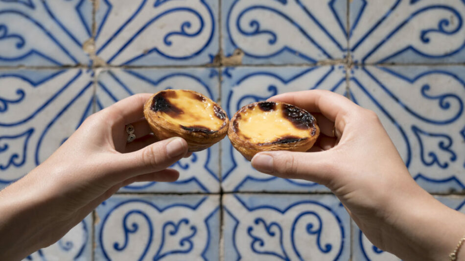 A pair of hands holding two pastries in front of a blue and white tiled wall in Portugal.