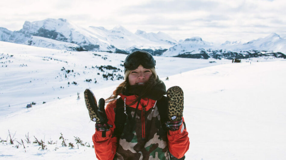 A woman embracing self love stands atop a snowy mountain in a camouflage jacket.