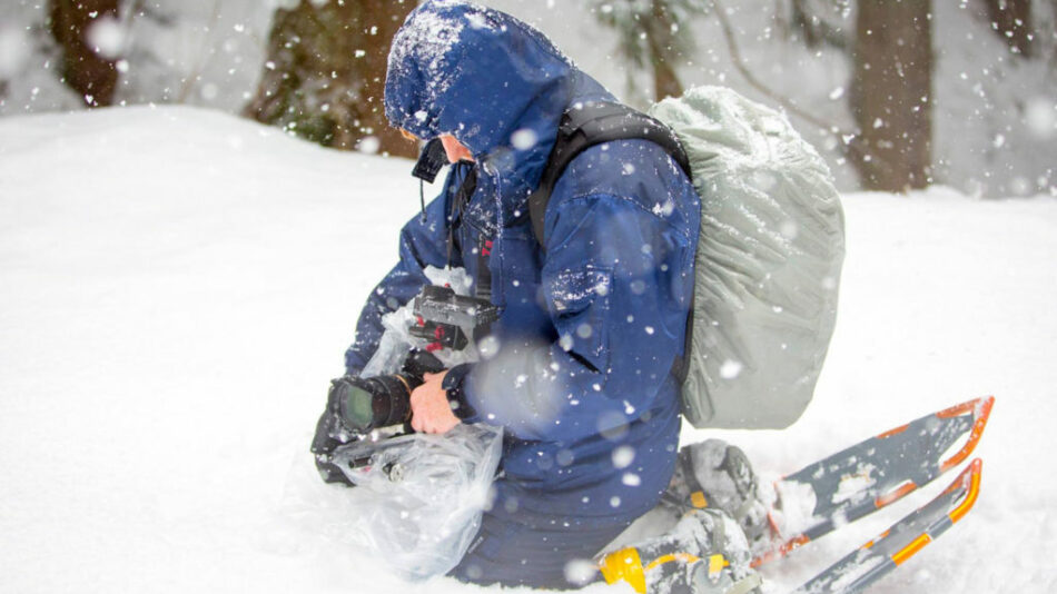 Travel filmmaker capturing a man kneeling in the snow with a backpack.