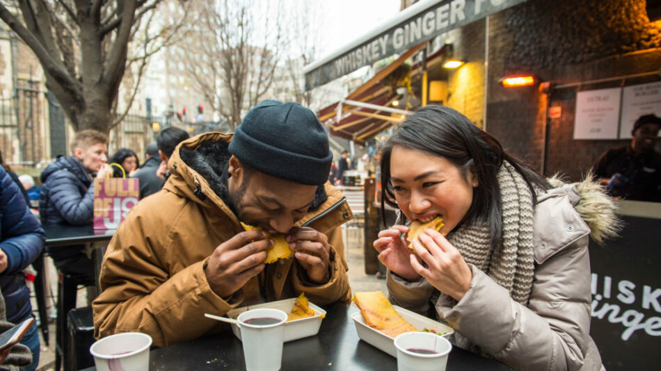 Two people eating food at an outdoor table in the UK.