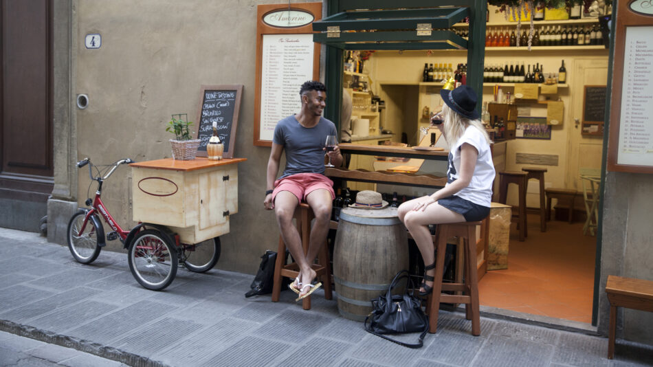 Two people enjoying Italian food while sitting on a bench in front of a wine bar.