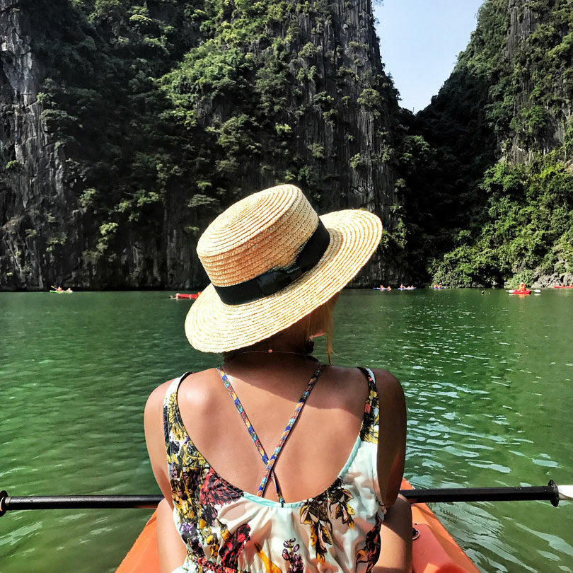 A woman in a straw hat is paddling a kayak on a lake while showcasing Hoi An fashion.