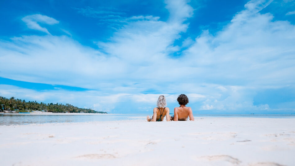 Two people sitting on a sandy beach under a blue sky in Thailand.