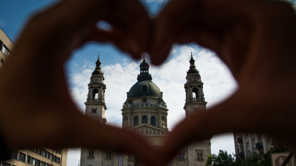 A person makes a heart shape with their hands in front of a church in the Balkans.