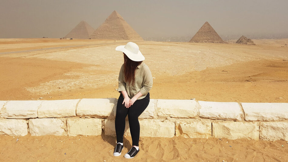 A woman sitting on a wall in front of the pyramids.