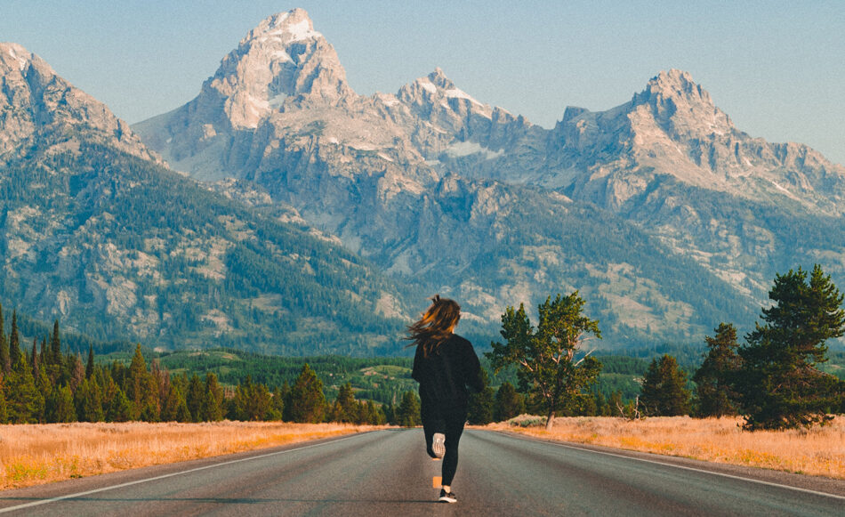 In one of the best places to visit in August, a woman is walking down a road mesmerized by the majestic mountain range in the background.