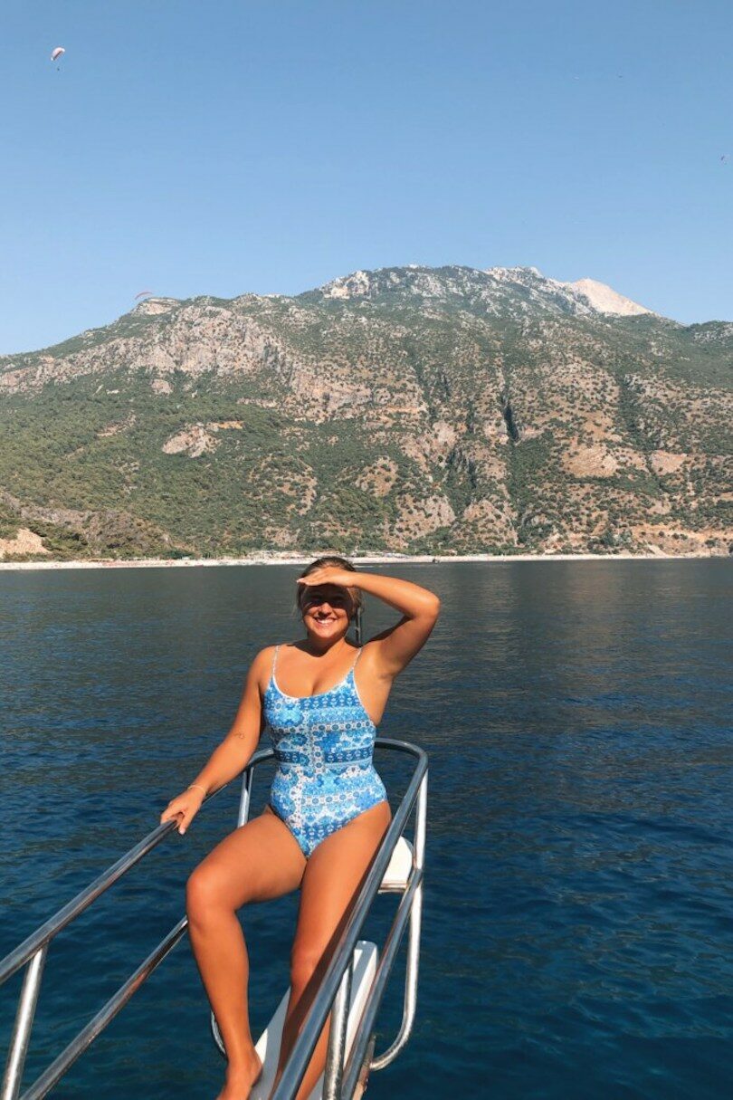 A woman in a blue swimsuit gracefully lounges on a boat while surrounded by the picturesque Turkish mountains in the background.