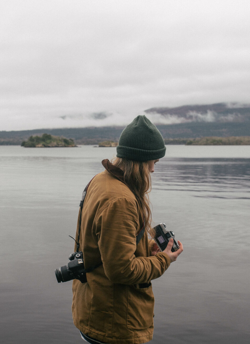 A woman of Irish roots holding a camera in front of a body of water.