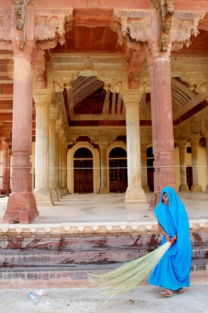 A posed woman in blue carries a broom in front of an ornate building.