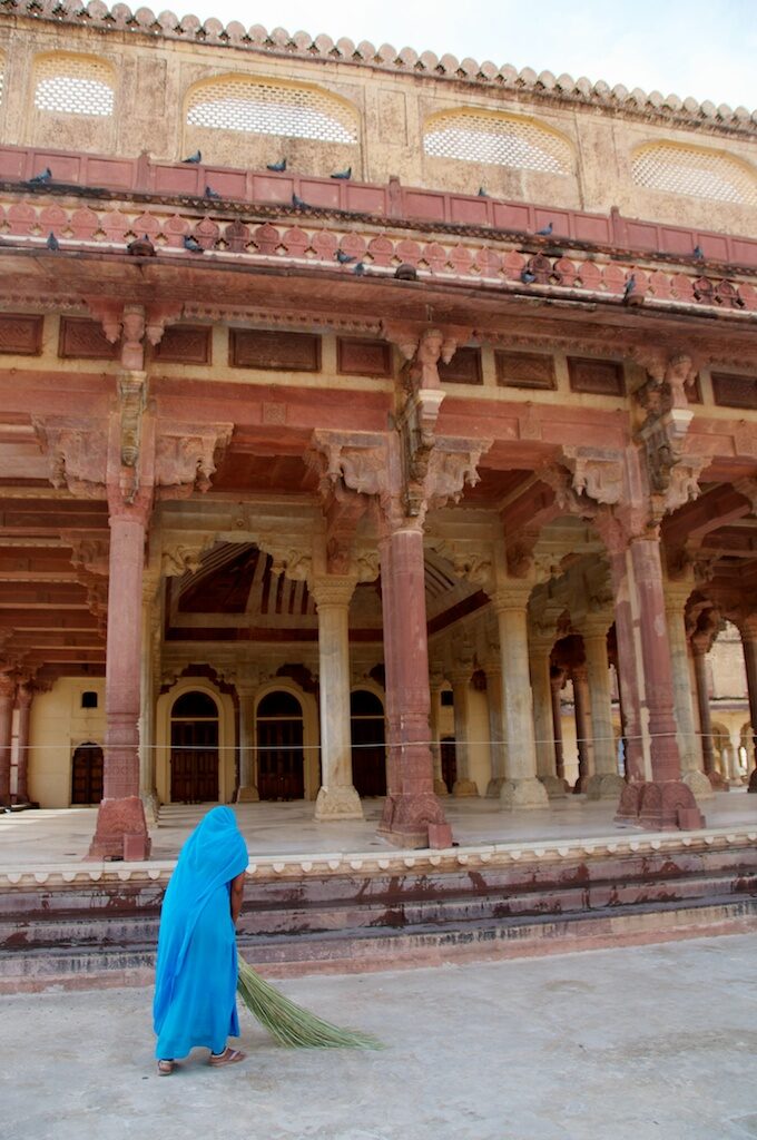 A posed woman in a blue sari is sweeping in front of an ornate building.