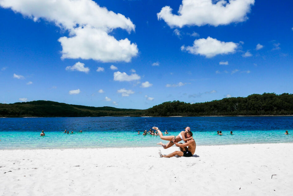 Two people sitting on a white sand beach near a body of water, capturing beautiful travel photos.