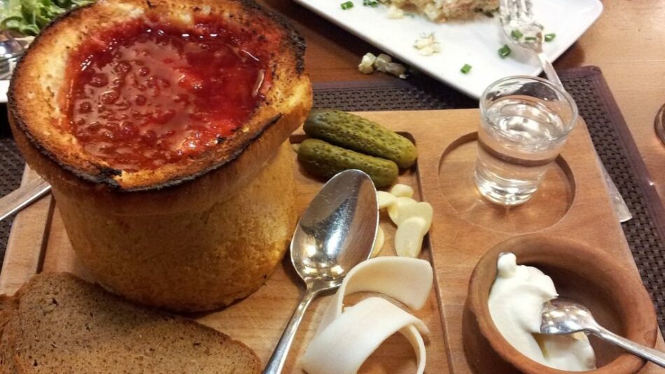 A bowl of soup with pickles and bread on a table.