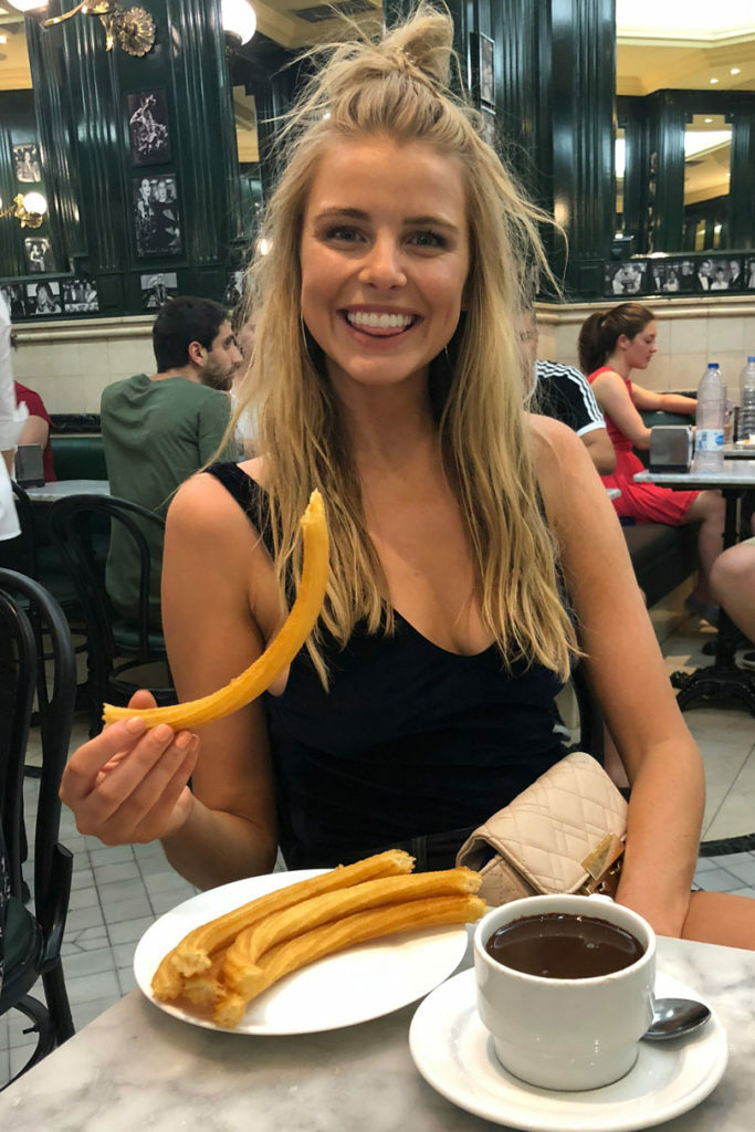 A blonde woman holding a plate of fried churros and a cup of coffee.