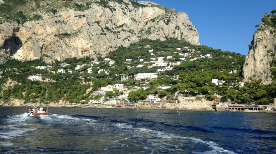 A boat is traveling down the water next to a mountain in Italy.