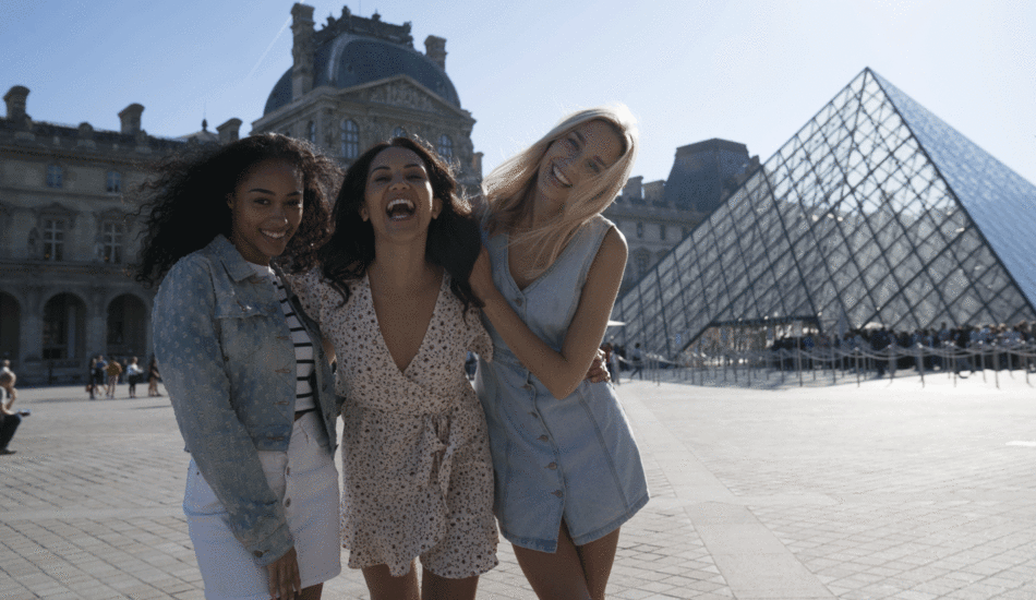 Three women posing in front of the Louvre pyramid, displaying social confidence.