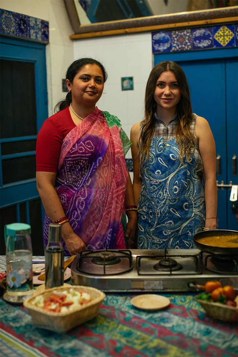 Two Indian women standing in front of a stove with food on it.